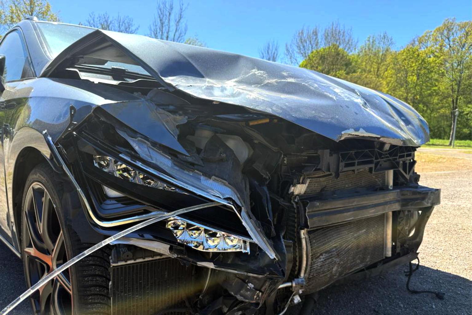 Ein verunfalltes Auto mit schwer beschädigter Frontpartie, zerbrochenem Scheinwerfer und deformierter Motorhaube, auf einer Straße bei sonnigem Wetter mit Bäumen im Hintergrund.