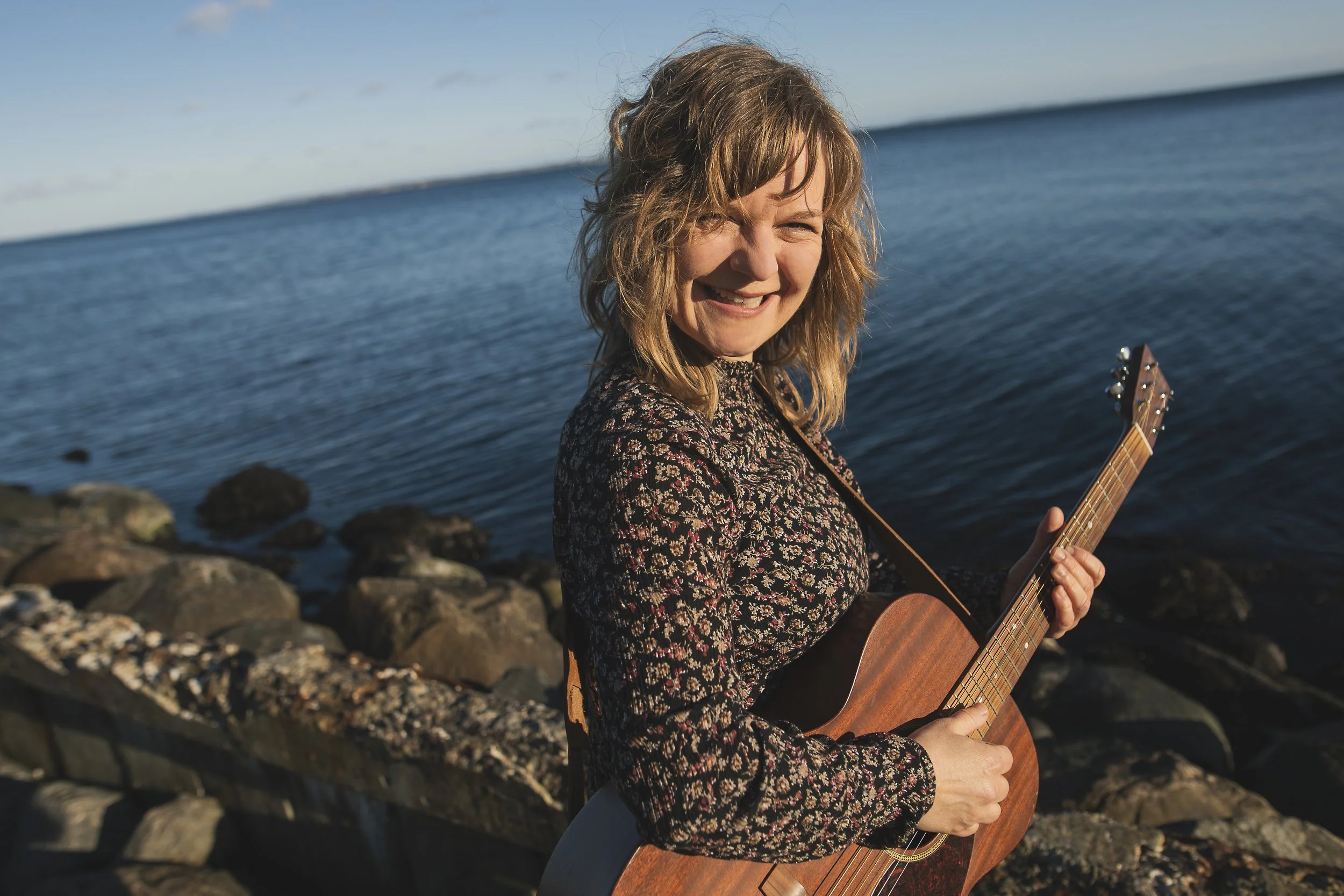 Kvinde med brunt, krøllet hår spiller på en guitar ved stranden med sten og hav i baggrunden.