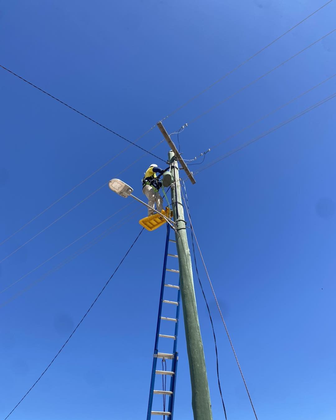 Utility worker repairing power lines on a wooden utility pole using a bucket ladder under a clear blue sky
