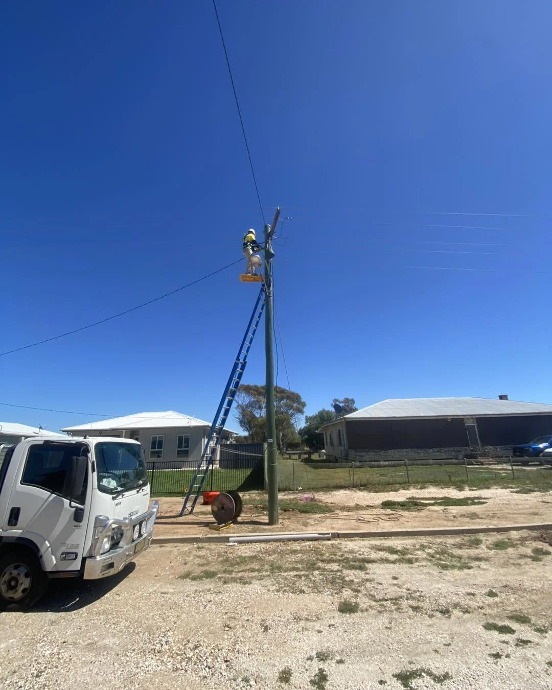 Utility worker repairing power lines from a ladder on a wooden utility pole beside a service truck in a residential neighborhood