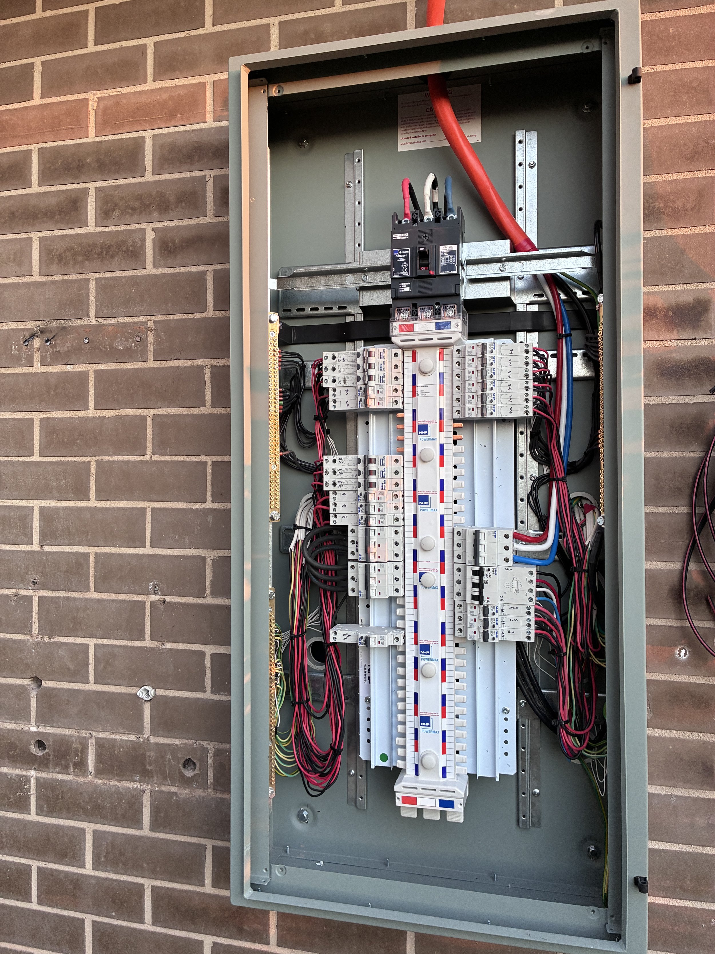 Open electrical distribution panel mounted on a brick wall, showing circuit breakers, busbars, wiring, and a large main breaker.