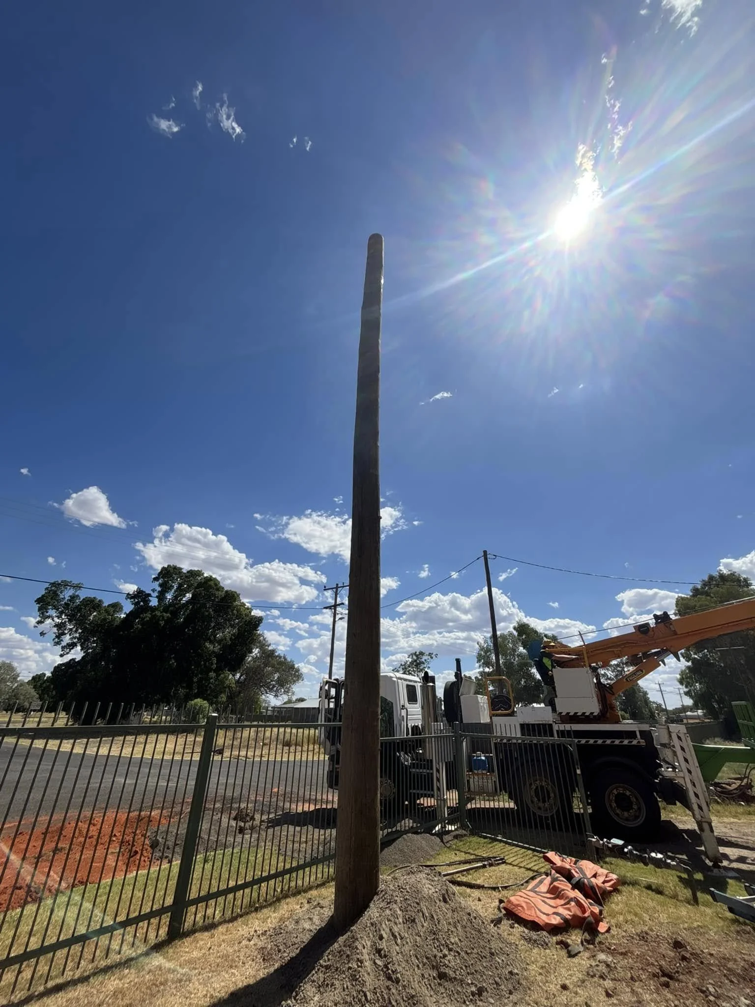 Utility crew installing a tall wooden utility pole beside a fenced property under a bright blue sky