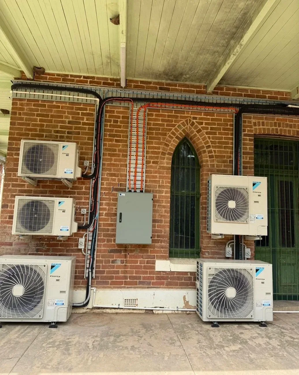 Five Daikin outdoor air-conditioning units mounted on a red brick building with exposed wiring and a central electrical panel