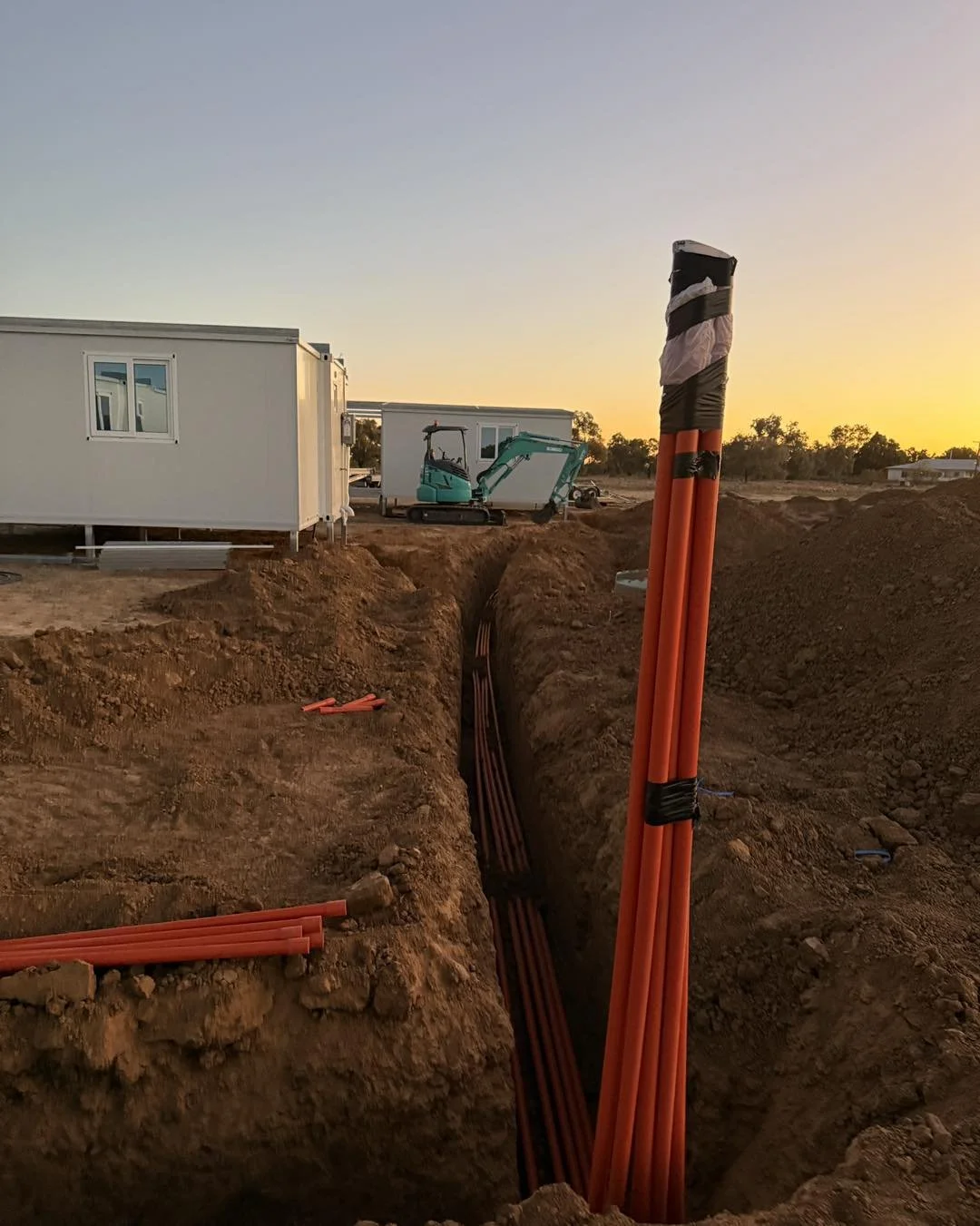 Construction site at sunset with orange utility conduits in a deep trench, excavator, and modular buildings