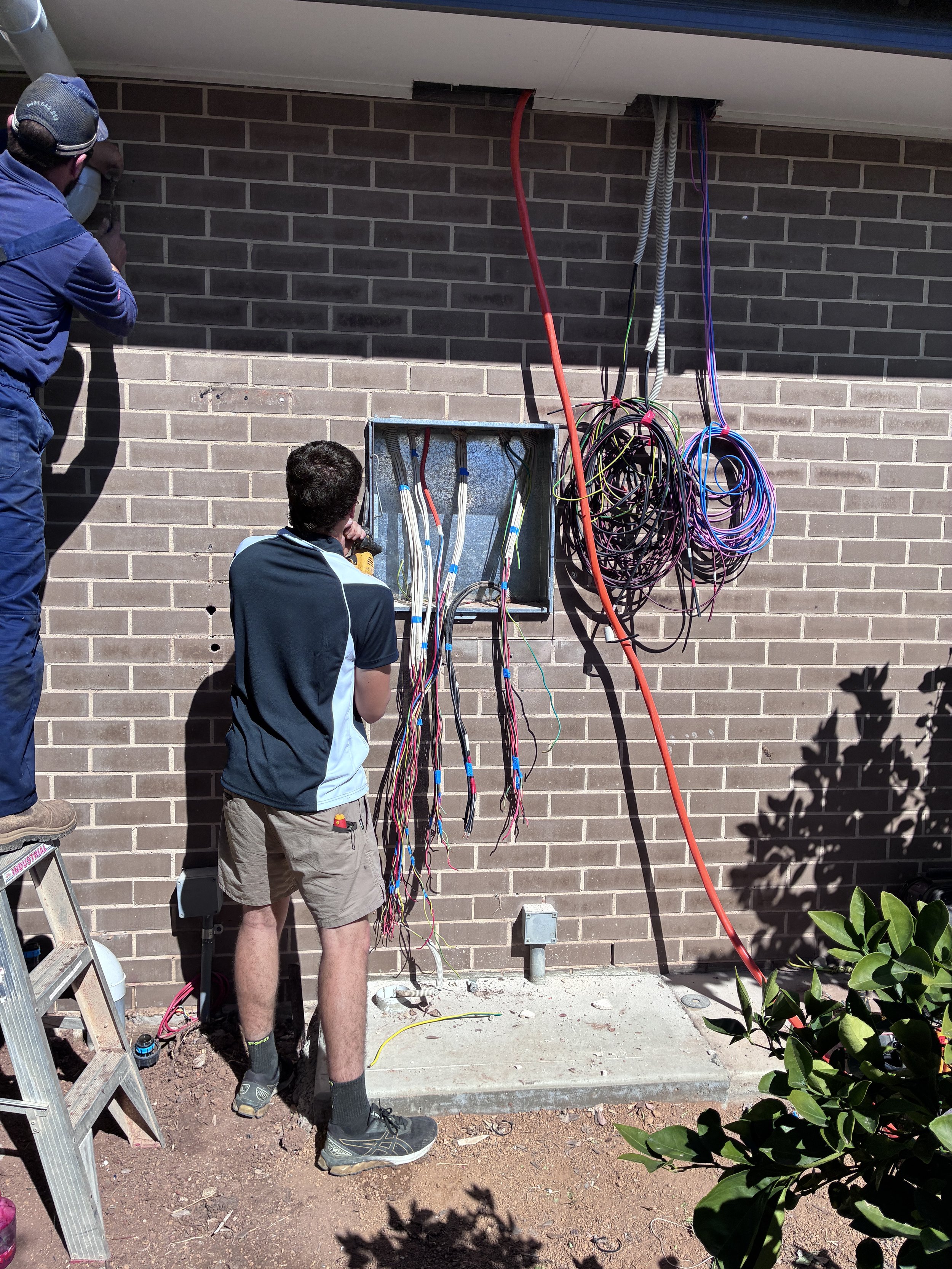 Technicians install electrical wiring and conduit in an open utility panel on a brick building.