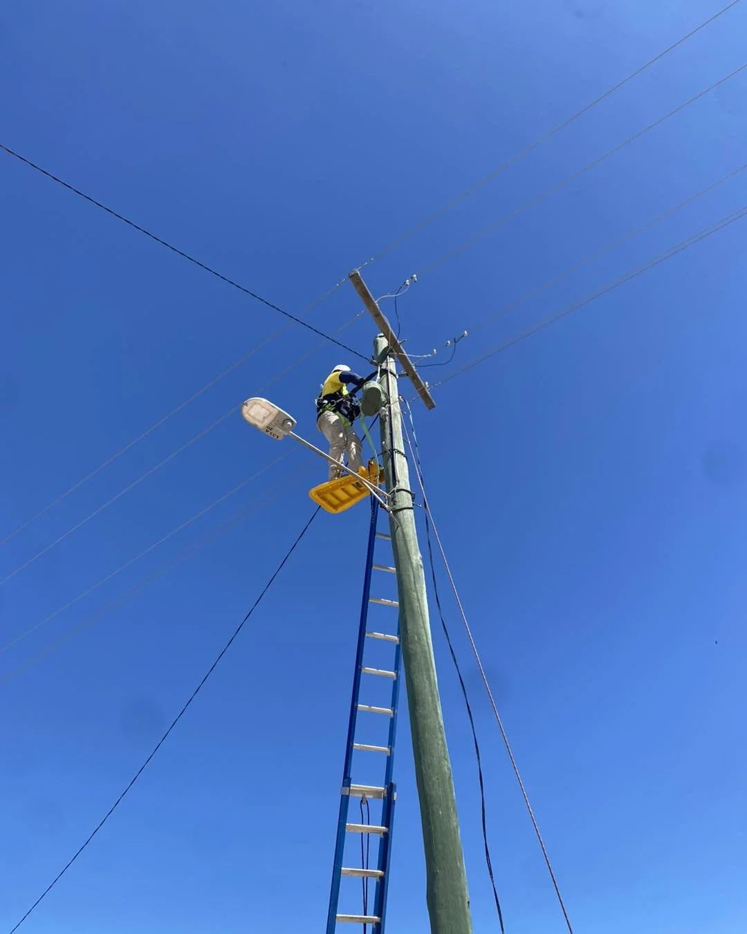 Utility worker repairing power lines on a wooden utility pole from a raised bucket under a clear blue sky