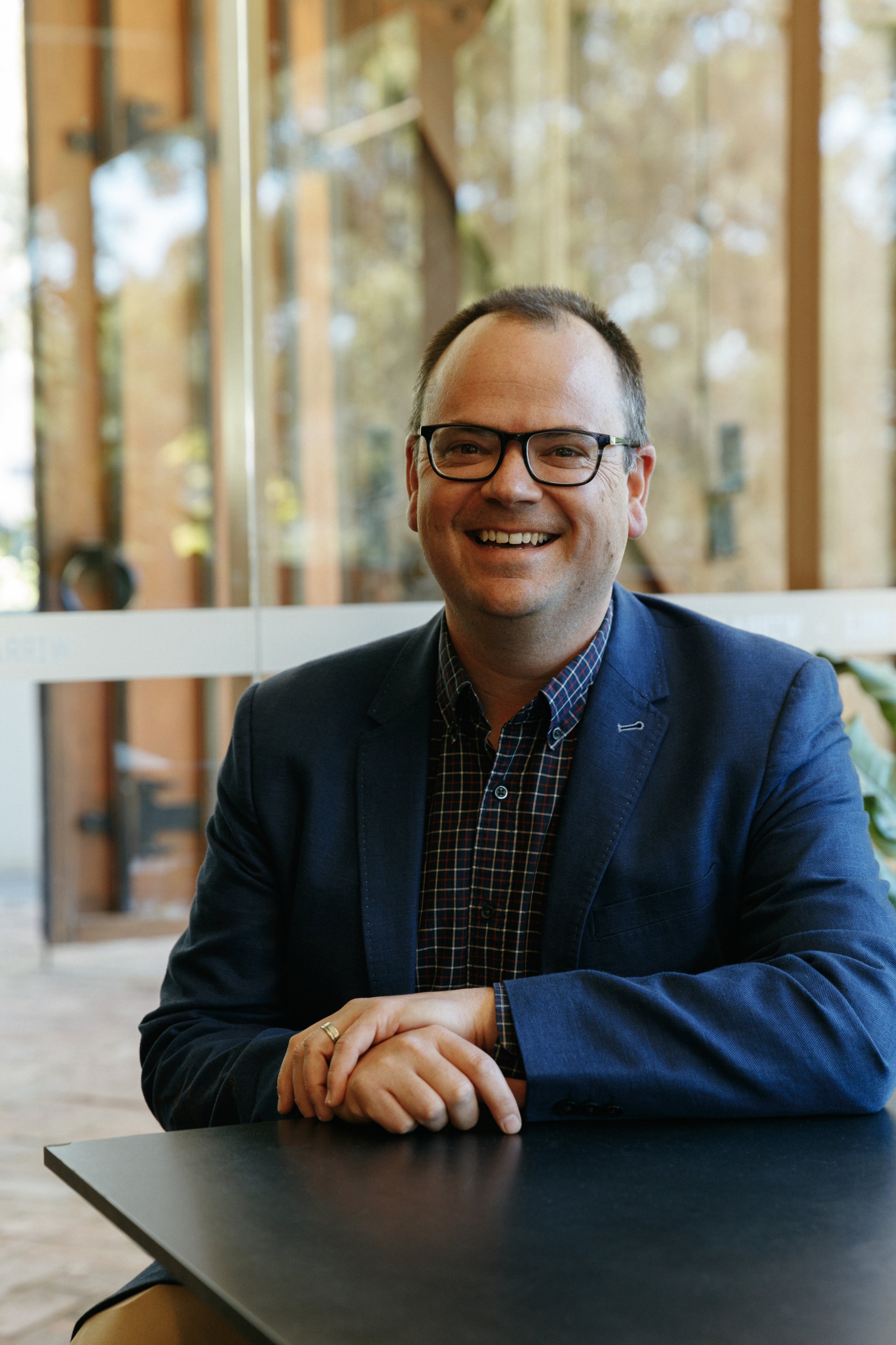 Portrait of a smiling man wearing glasses, a dark blue blazer, and a checkered shirt, sitting at a table in a modern office or conference room with large windows and a wooden interior visible in the background.