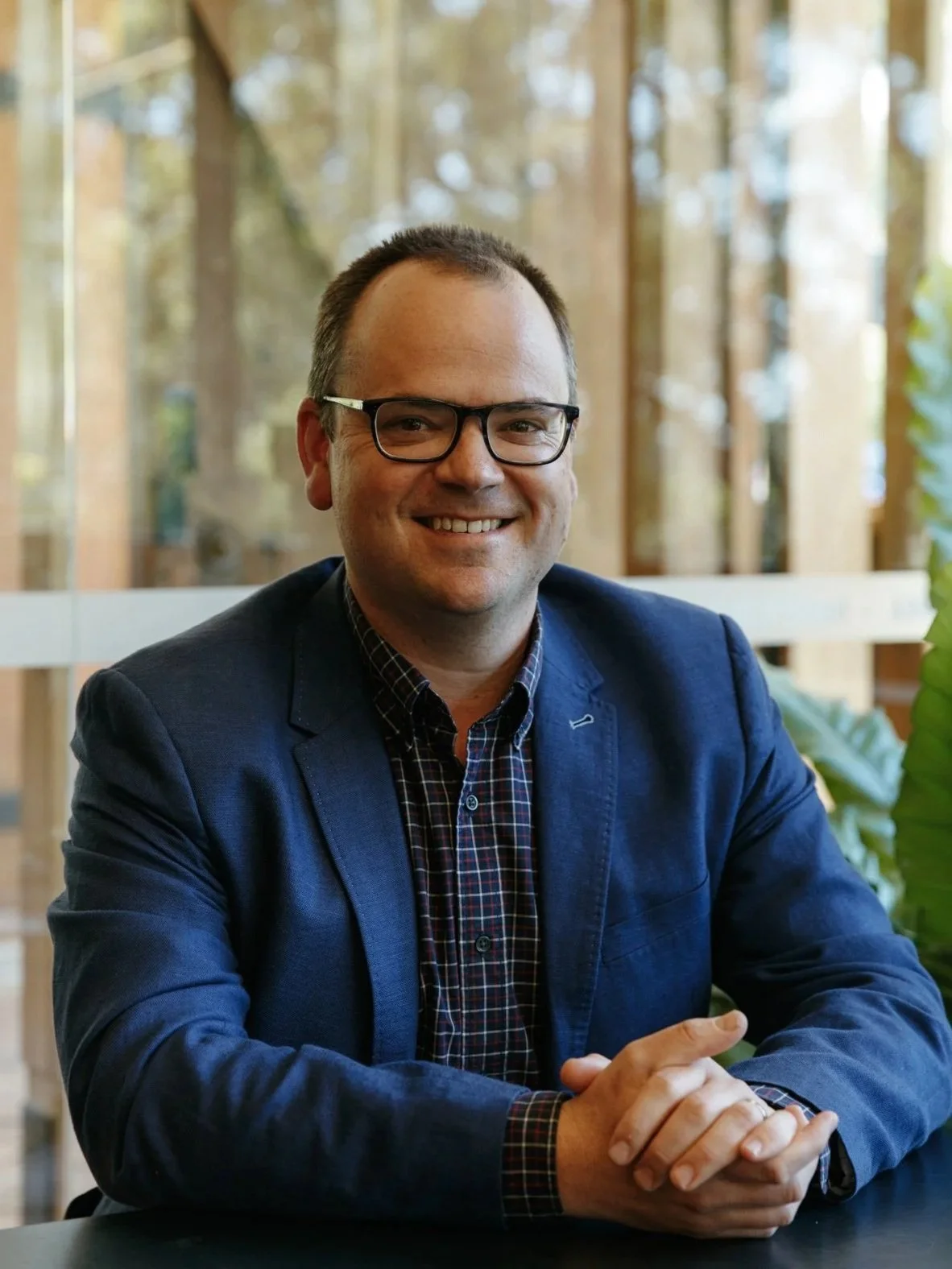 Smiling man with glasses wearing a dark blazer and a checkered shirt, sitting at a table in a bright room with large windows and greenery outside.