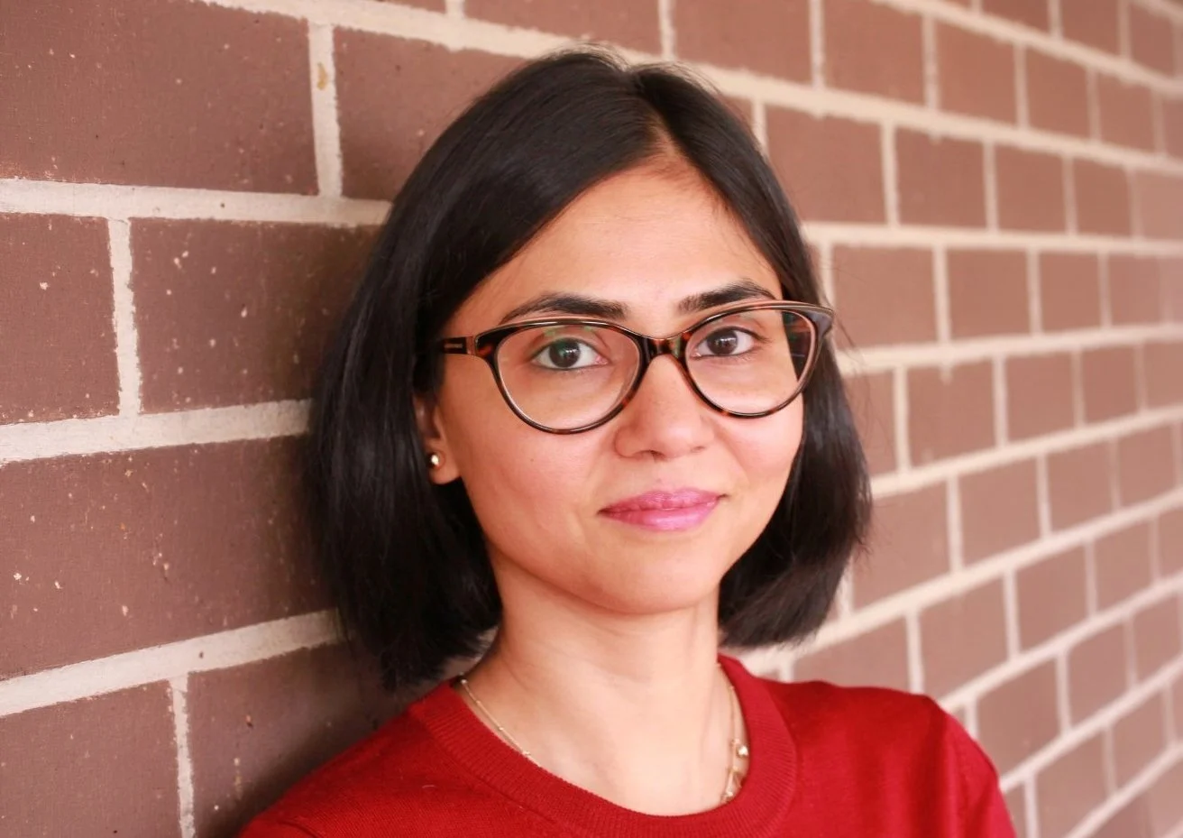 A woman with short black hair and glasses standing against a brick wall, wearing a red top and gold jewelry.