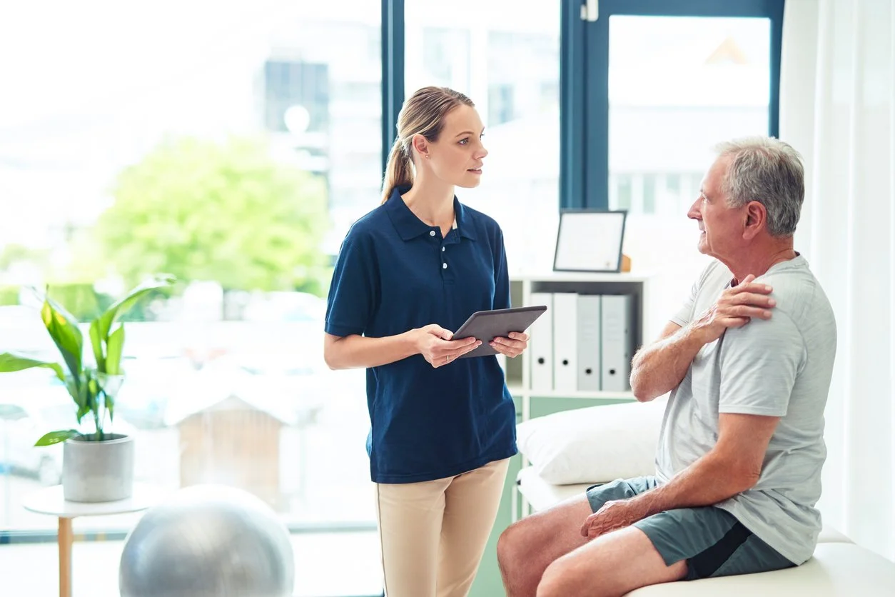 A healthcare professional talking to an older man in a medical office, with the man sitting on an examination table.