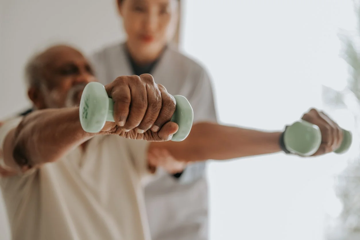 An elderly person and a caregiver are exercising together, holding small green dumbbells.