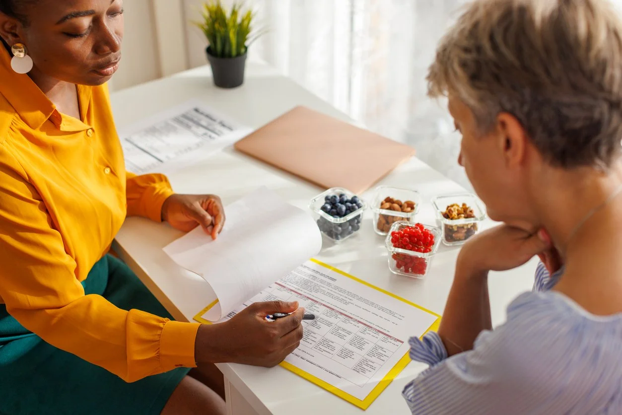 Two women sitting at a white table discussing a document, with fruit containers and a laptop on the table.