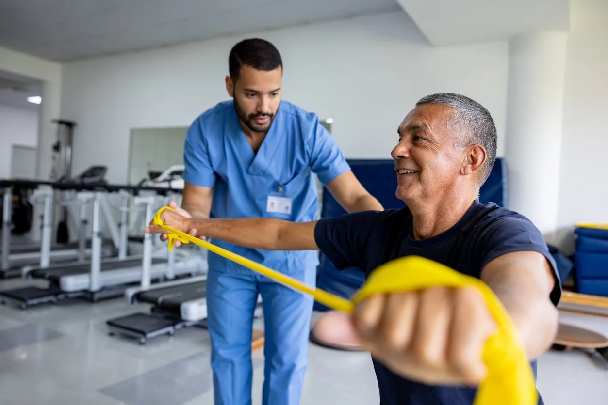 A healthcare worker is assisting an elderly man with resistance band exercises in a rehabilitation setting.