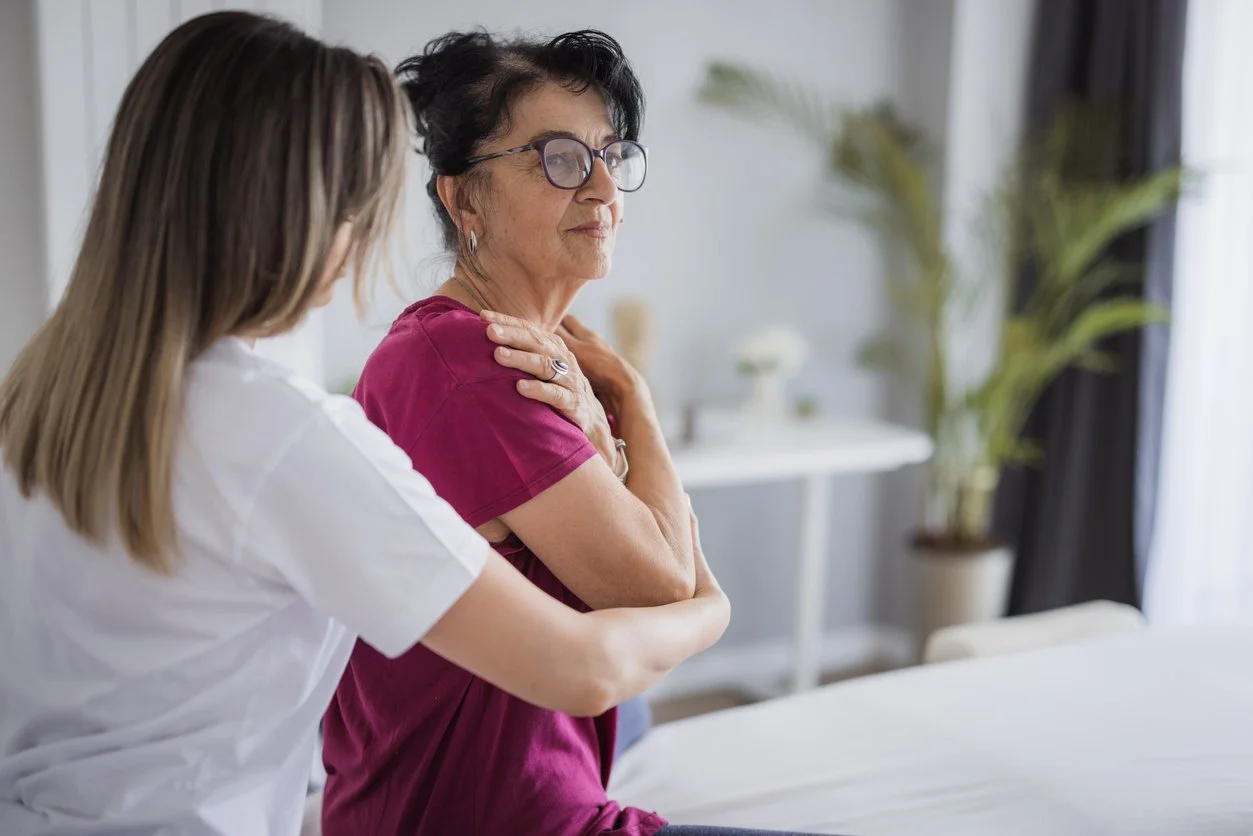 A healthcare professional attends to an elderly woman in a medical setting, with the woman holding her shoulder.