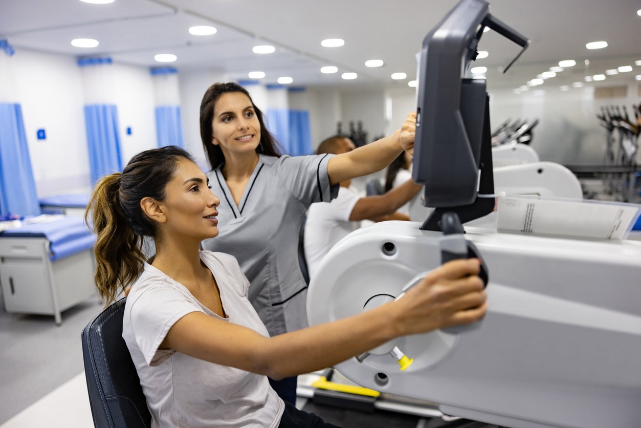 Medical training in a hospital room, one woman sitting at a medical simulator and another woman instructing, with other medical trainees in the background.