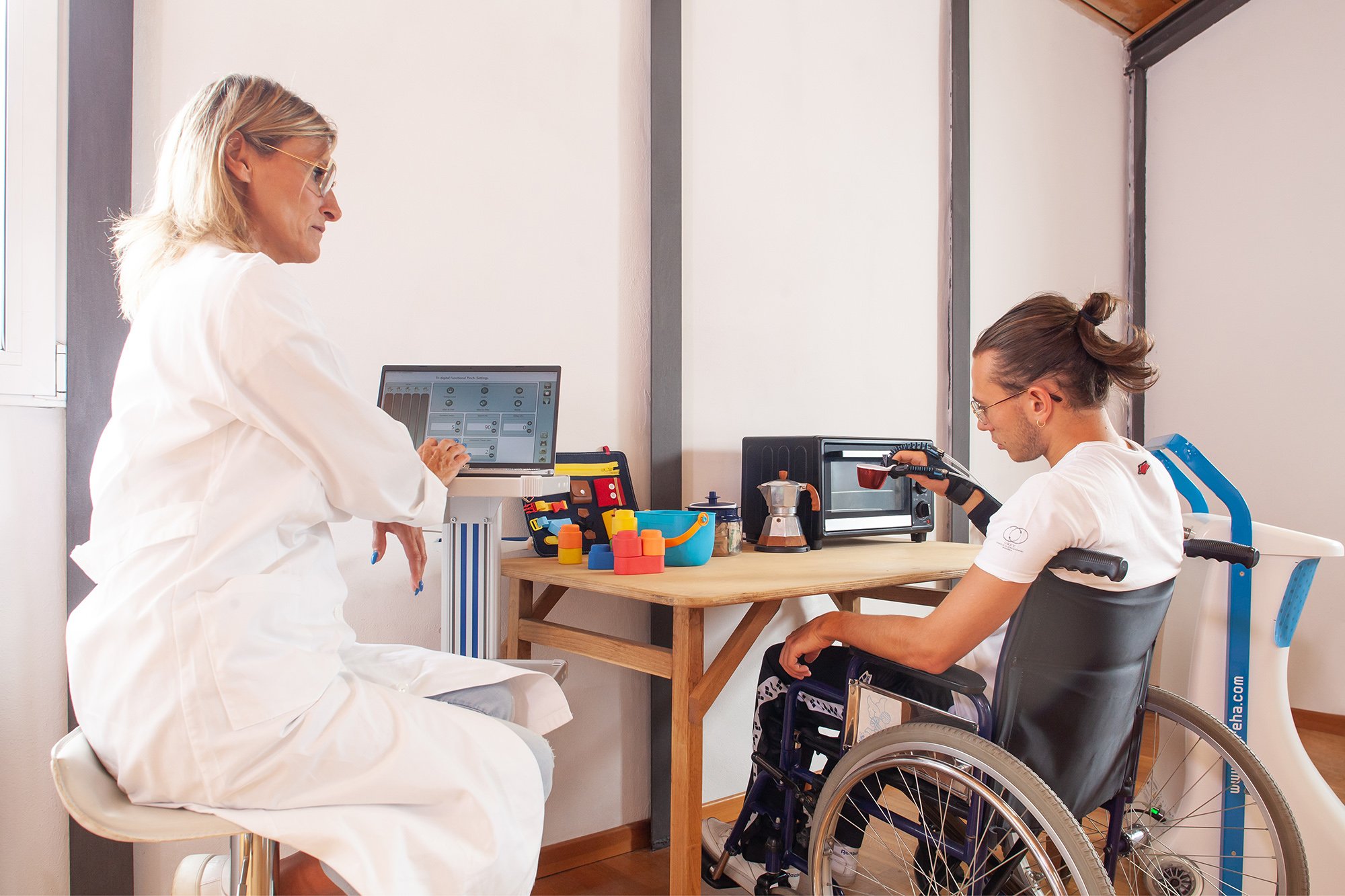A healthcare professional in a white coat talking to a man in a wheelchair in a clinical setting with medical equipment and toys on the table.