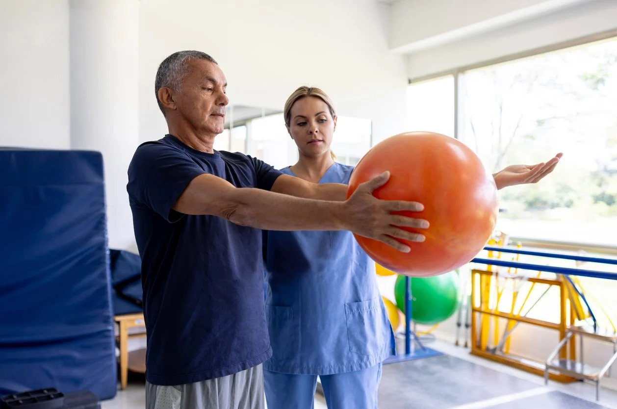 A senior man in a navy shirt practicing balance with a therapy ball guided by a female healthcare professional in a physical therapy clinic.