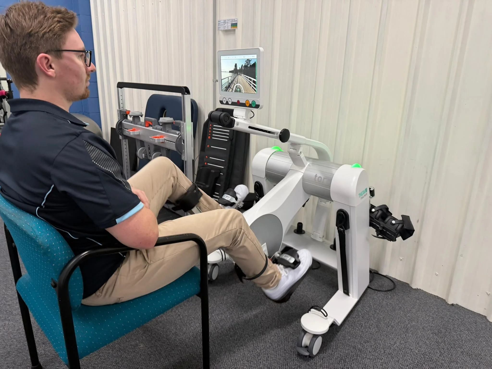 A man sitting in a blue chair using a rehabilitation robotic device with a screen displaying a virtual outdoor scene.