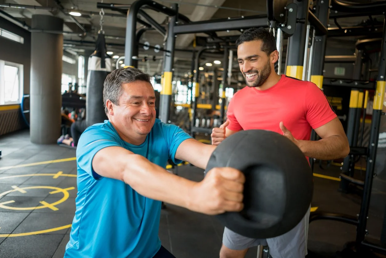 Two men working out with a large black weight in a gym, one lifting and the other spotting, both smiling.