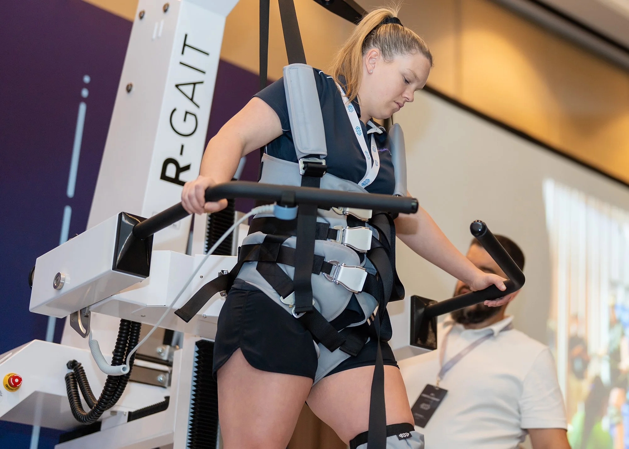 A young woman in athletic attire and a harness is walking on a treadmill with support from a harness system, likely during a physical therapy or rehabilitation session, with a man standing nearby.