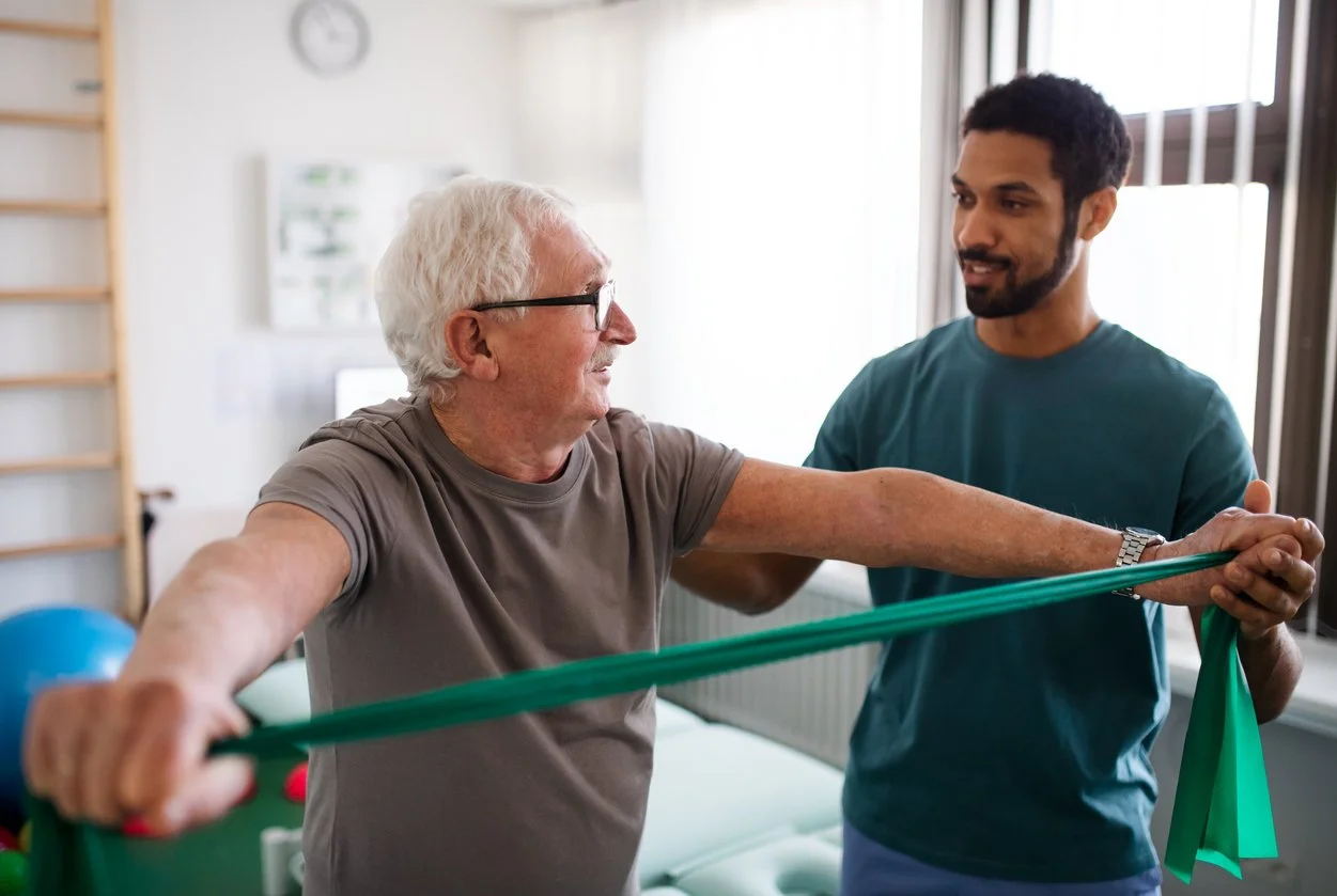 A male elderly patient doing physical therapy exercises with a resistance band at a rehabilitation center, assisted by a physical therapist.