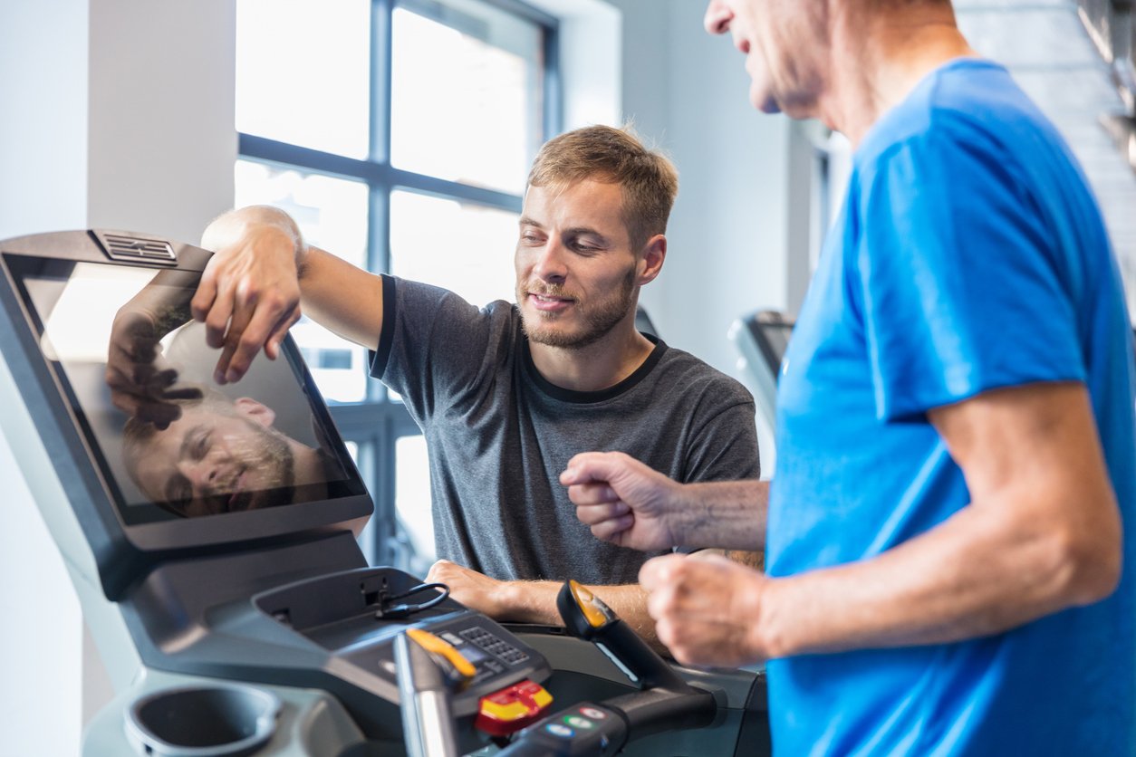A young man and an older man are at the gym, with the young man standing next to a treadmill and the older man operating the treadmill controls. The young man is smiling and leaning on the treadmill, while the older man, wearing a blue shirt, is looking at him. The gym has large windows letting in natural light.