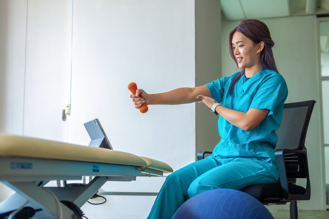 Healthcare worker in scrubs performing shoulder exercises with a small orange dumbbell in an office or clinic setting.