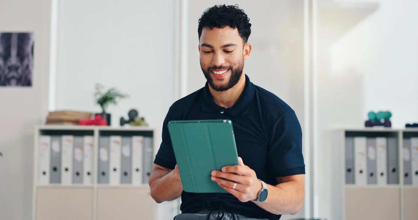 A man with dark curly hair, beard, wearing a navy blue polo shirt, smiling while looking at a tablet in an office setting with binders and plants in the background.