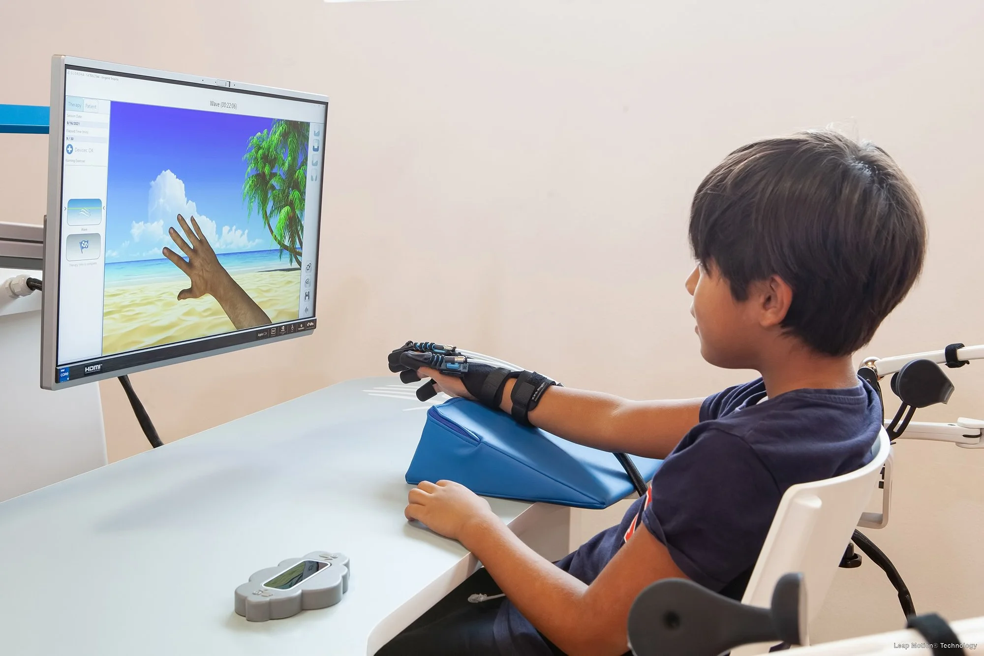A young boy seated in a white chair during a medical or therapy session is touching a computer screen displaying a virtual beach scene with a tree, blue sky, and ocean. The boy's right arm is extended and resting on a blue cushion on the table in front of him, with a medical device on his wrist and a handheld controller nearby.