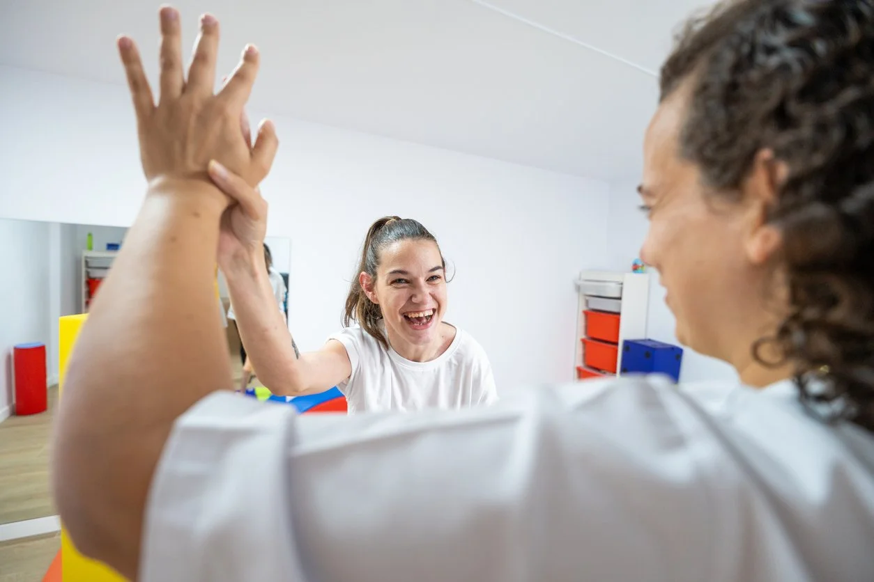 Two women are practicing hand therapy exercises, with one woman smiling and the other assisting in a brightly-lit room with colorful storage units in the background.