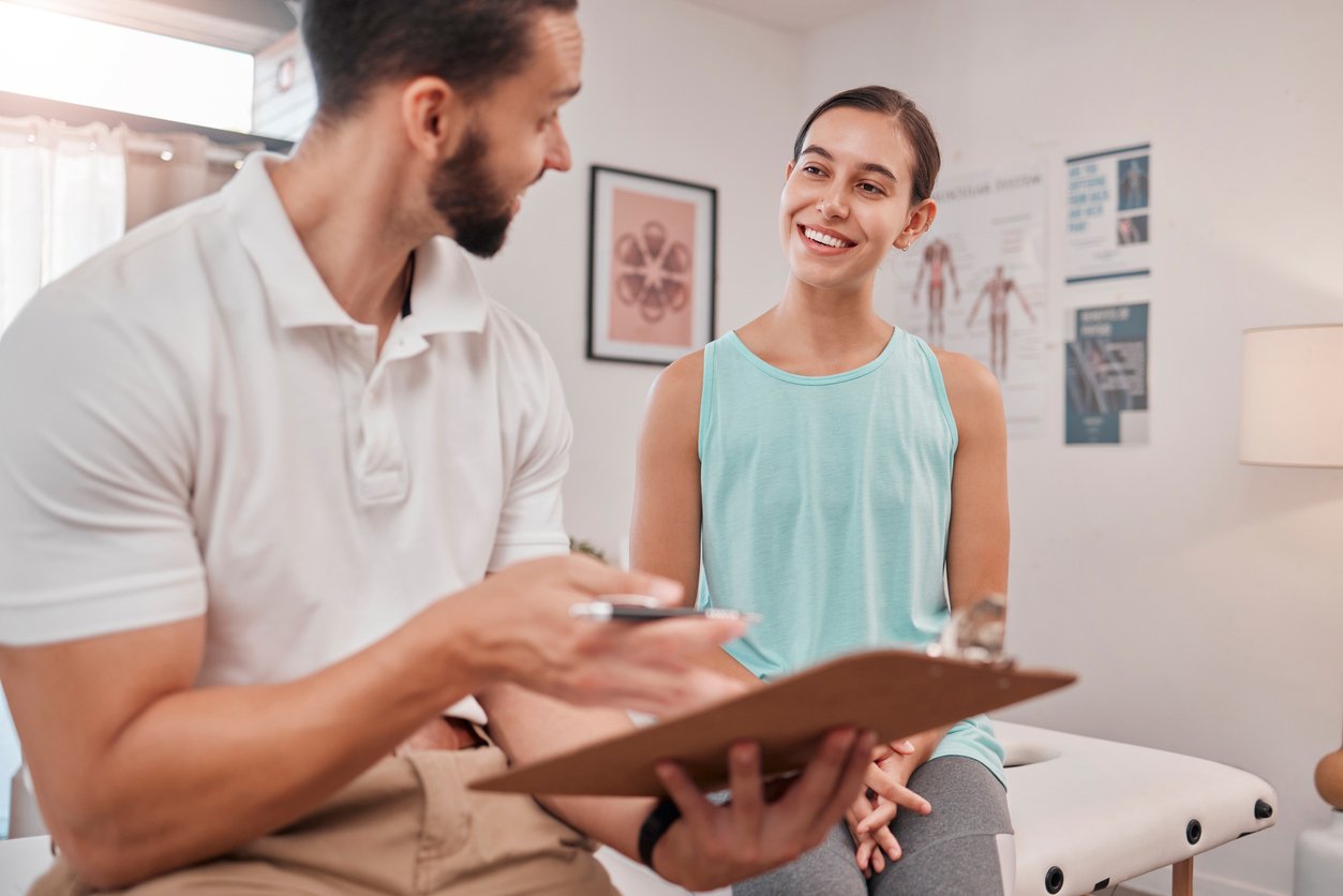 A woman smiling at a man sitting with a clipboard in a medical examination room.