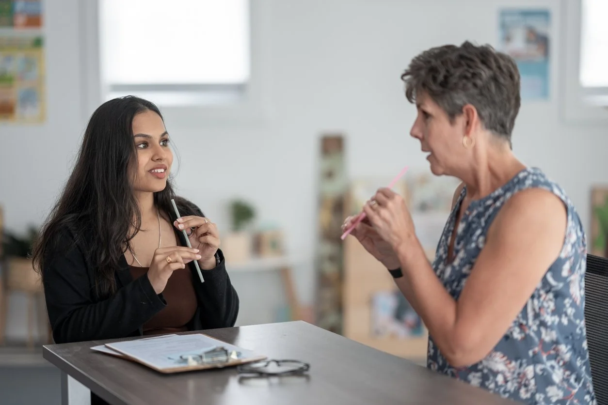 A young woman and an older woman are having a conversation at a table in an office or classroom setting. The young woman has long dark hair and is holding a pen, while the older woman is speaking animatedly, also holding a pen.
