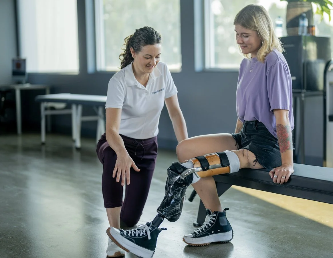 A woman with a robotic prosthetic leg sitting on an examination table while a female healthcare professional stands nearby, smiling indoors with large windows in the background.