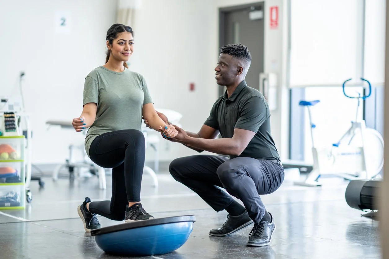 A woman performing balance exercise on a Bosu ball assisted by a male trainer in a rehabilitation or fitness center.