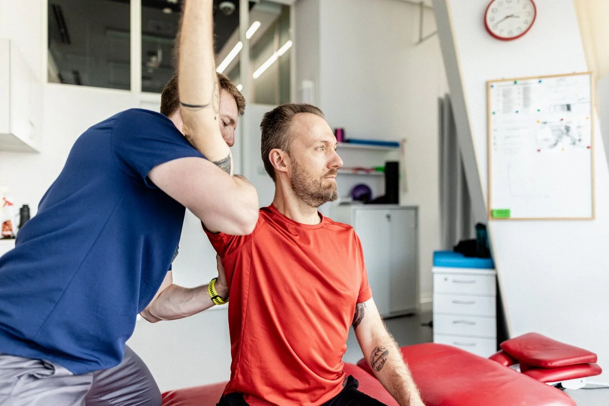 Physical therapist assisting a man in a red shirt with shoulder exercises in a clinic.