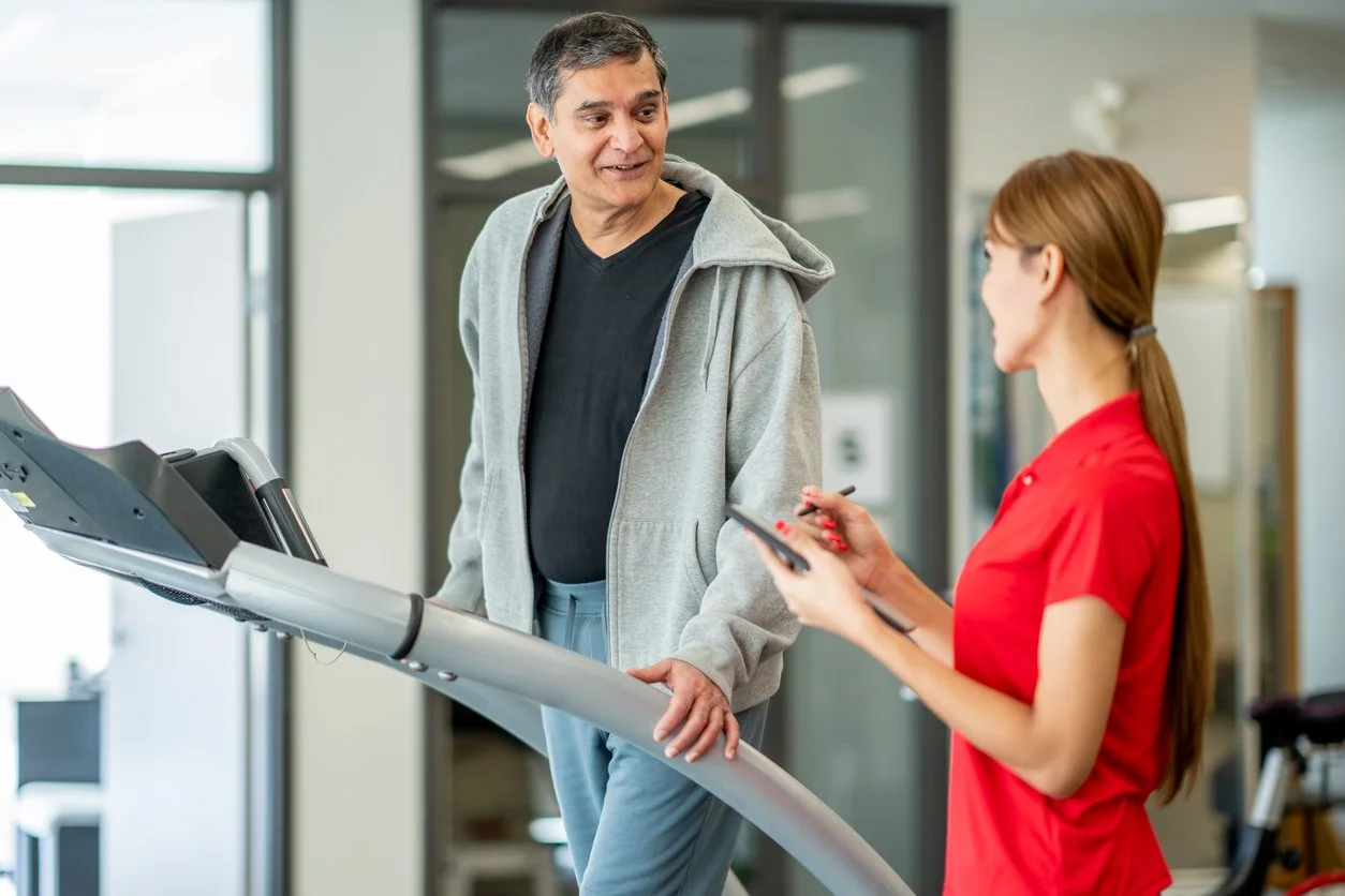 A middle-aged man in a gray hoodie and black shirt talking to a woman in a red shirt on a treadmill in a gym.