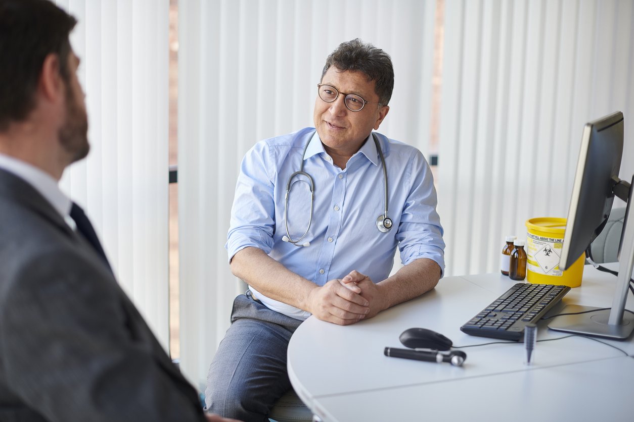 Male doctor wearing glasses and a stethoscope talking to a male patient in an office with medical supplies on the desk.