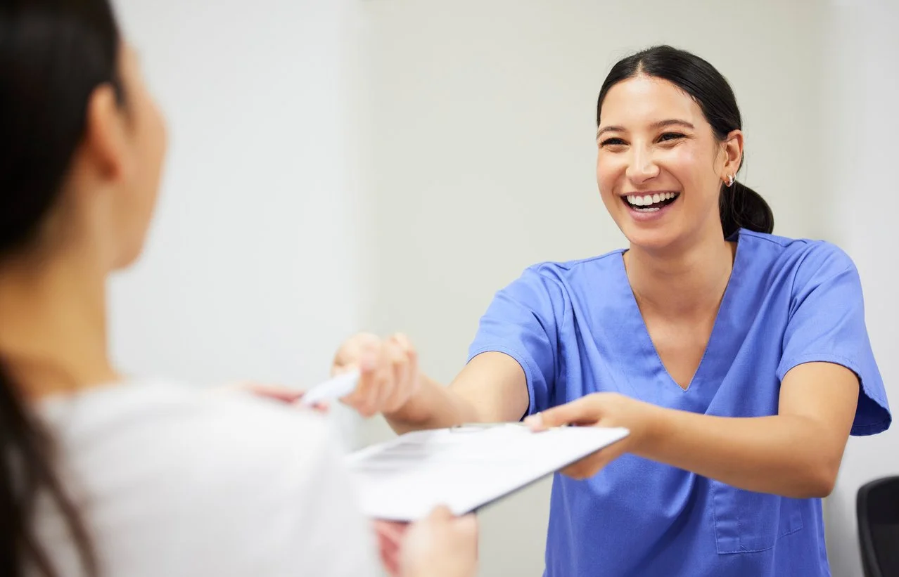 A smiling female healthcare worker in blue scrubs handing a clipboard to a patient in a clinical setting.
