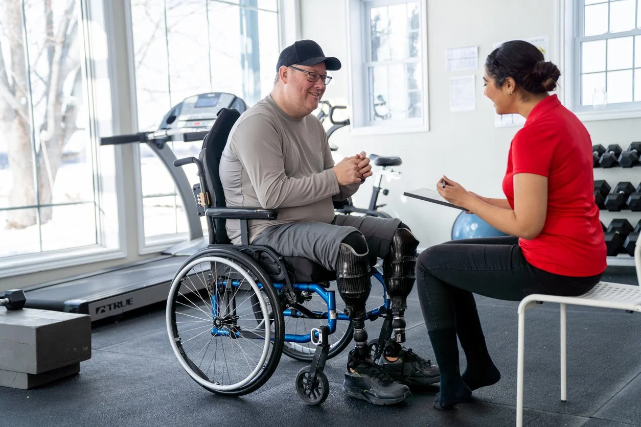 A person in a wheelchair talking to a woman with notepad in a gym with exercise equipment and large windows.