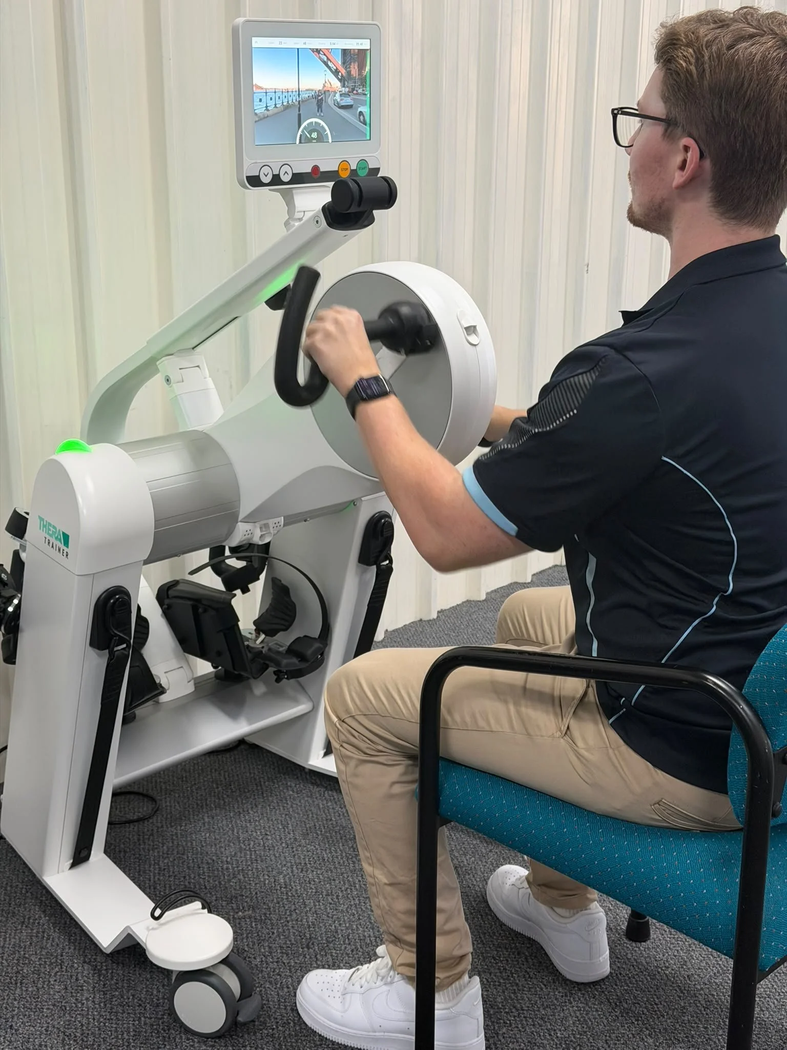 A person sitting in a wheelchair using a robotic exoskeleton device for rehabilitation therapy, with a monitor displaying a virtual walking scene.