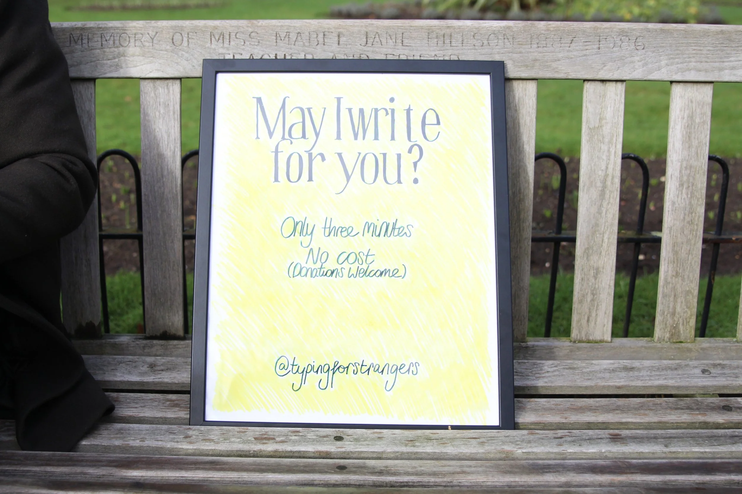 A sign on a yellow background reads 'May I write for you?' with smaller text underneath stating 'Only three minutes, no cost, donations welcome,' and a social media handle '@typingforstrangers'. The sign is placed on a wooden bench with a green park or garden background.