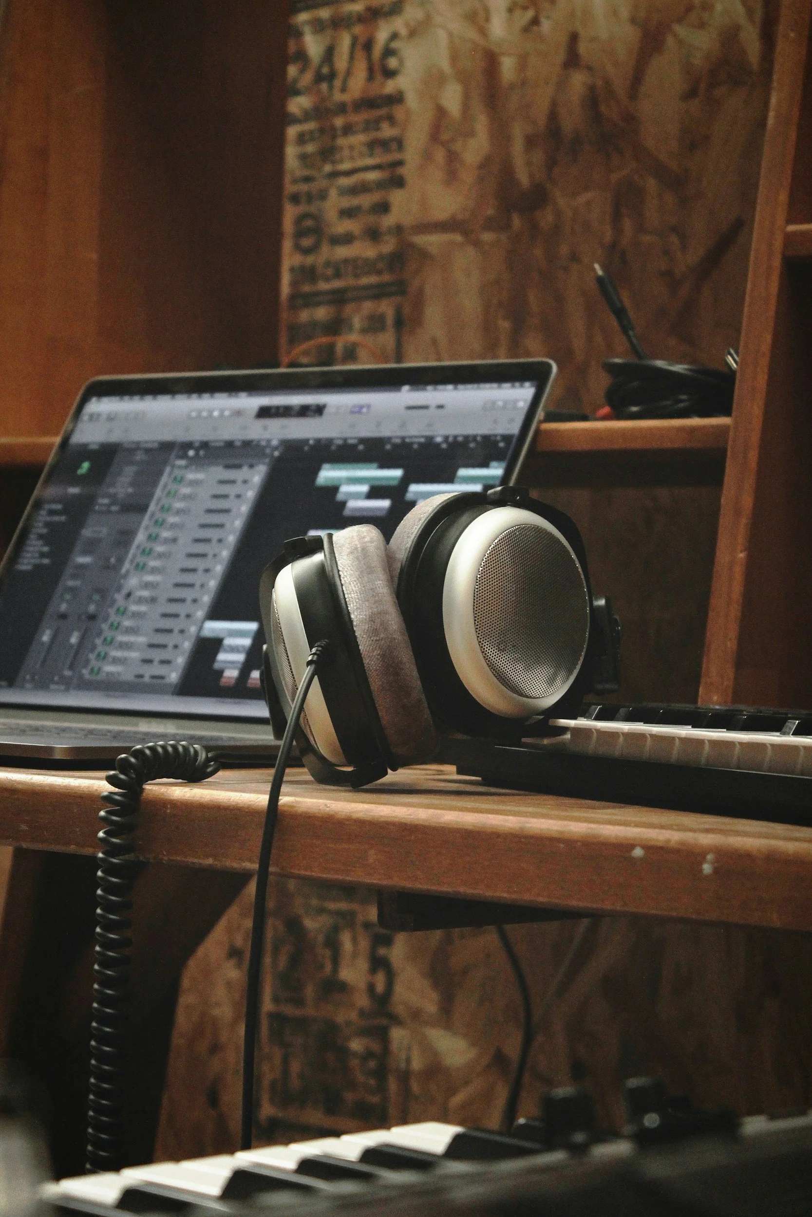 A music studio workspace featuring a laptop, a pair of headphones, and a keyboard, set against a wooden shelf with some electronics and a decorative wall background.