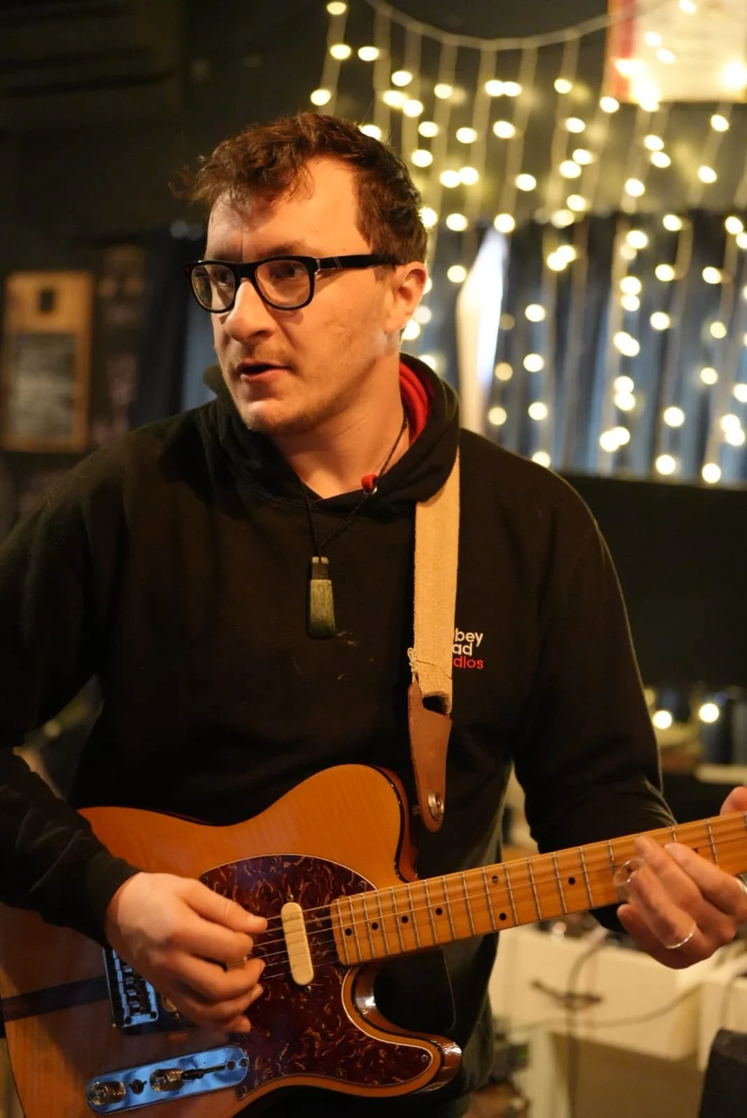 A man wearing glasses and a black hoodie is playing an acoustic guitar with a decorative pickguard. He is in a dimly lit room with string lights in the background.