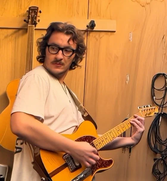 A man with curly hair, glasses, and a mustache playing an electric guitar in a recording studio.