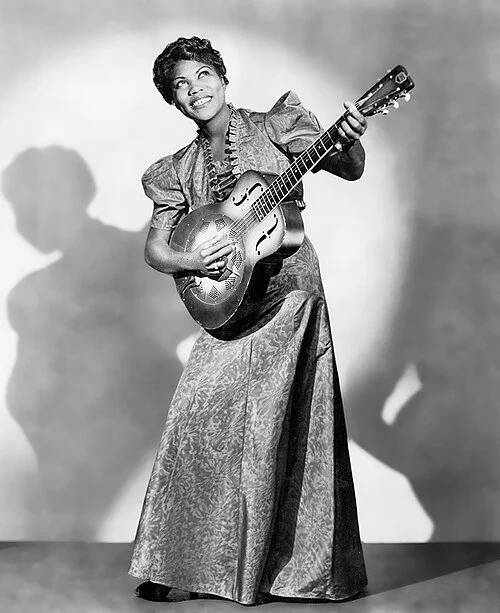 Black and white photo of a woman playing an acoustic guitar, smiling, with shadows on the wall behind her.