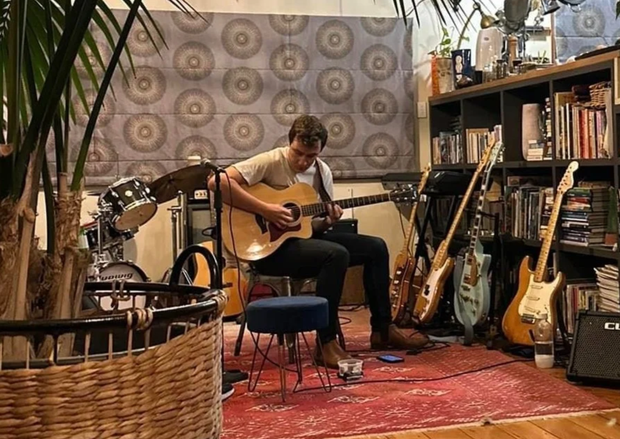 A young man playing an acoustic guitar in a cozy, decorated room with various musical instruments, bookshelves, and houseplants.