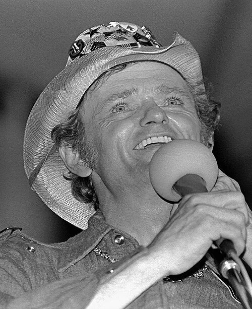 A man wearing a cowboy hat with a patterned band, smiling and holding a microphone close to his mouth as if singing or speaking.