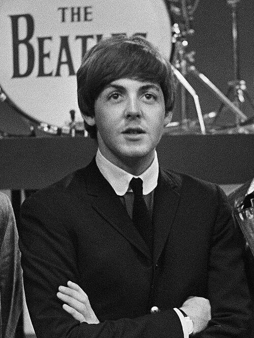 Black and white photo of a young man in a suit with arms crossed, standing in front of a drum set with 'The Beatles' logo on the bass drum.