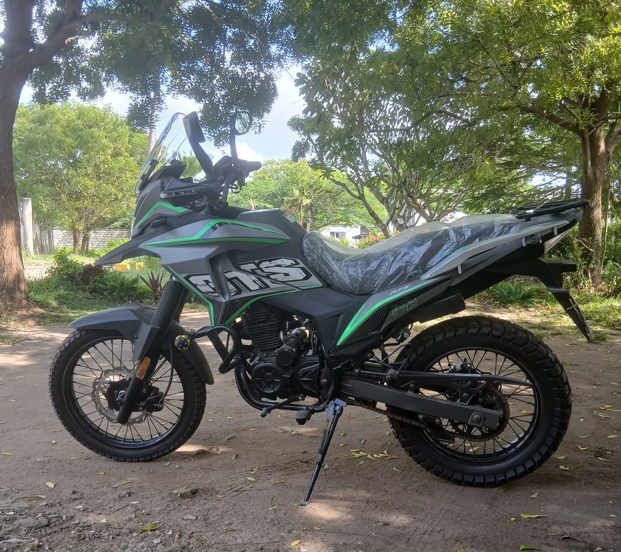 A black and green off-road motorcycle parked on a dirt path under a tree with lush green leaves. The motorcycle has a windshield, rugged tires, and a plastic bag covering the seat. There are additional trees and a fence in the background.