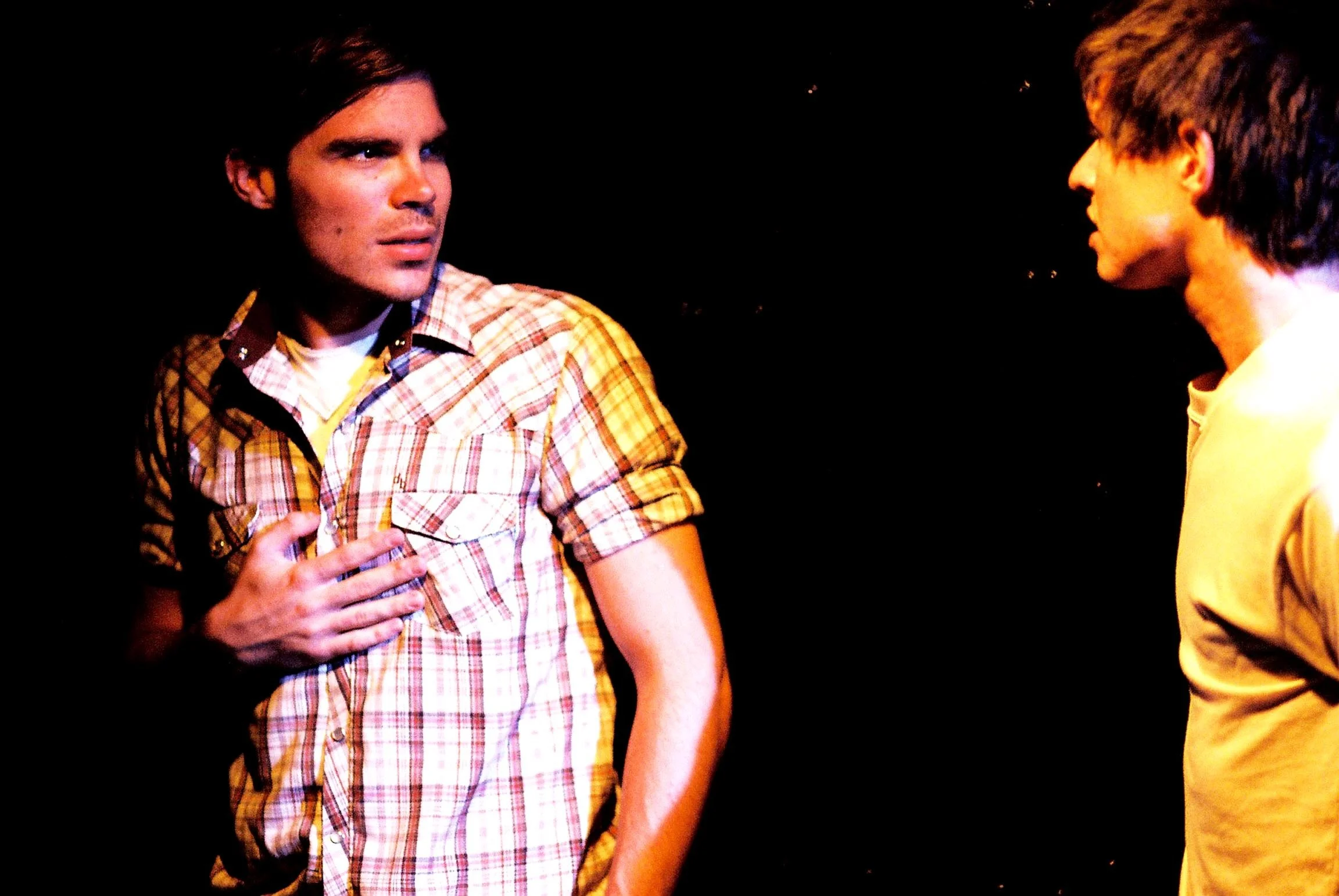 Two young men having a conversation in a dark room, one wearing a plaid shirt and the other wearing a yellow hoodie.